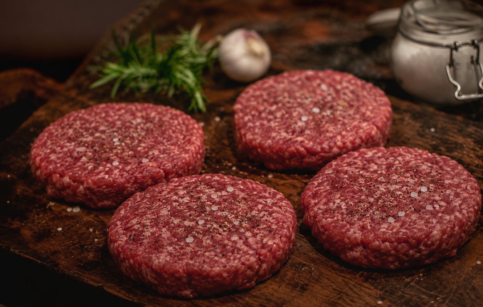 Vier Angus-Burger-Patties aus grob gewolftem Rindfleisch, mit Salz und Pfeffer gewürzt, auf dunklem Holzbrett