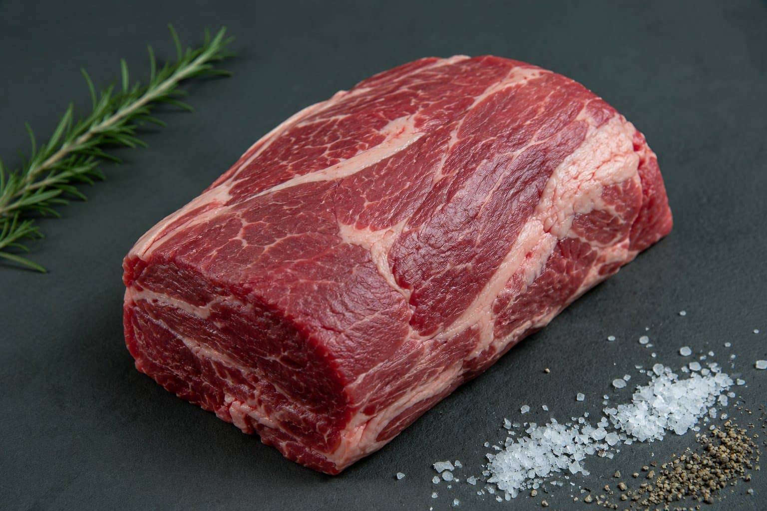 Schmorbraten vom Aberdeen Angus, gut durchwachsenes Stück auf dunkler Fläche mit Rosmarin, Meersalz und Pfefferkörnern