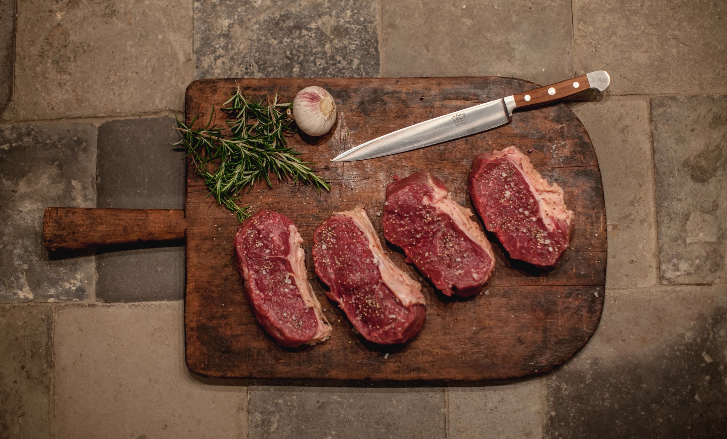 Vier Dry-Aged Steaks auf Holzbrett mit Messer und Rosmarin